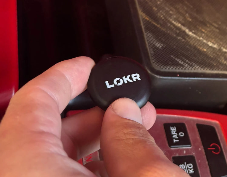 A close-up shot of a hand holding a LOKR tracking tag with white branding text, positioned above a digital scale