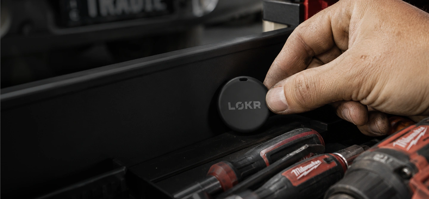 A close-up shot shows a hand placing a small, round, black tracking tag onto the inside wall of a dark-colored toolbox or drawer