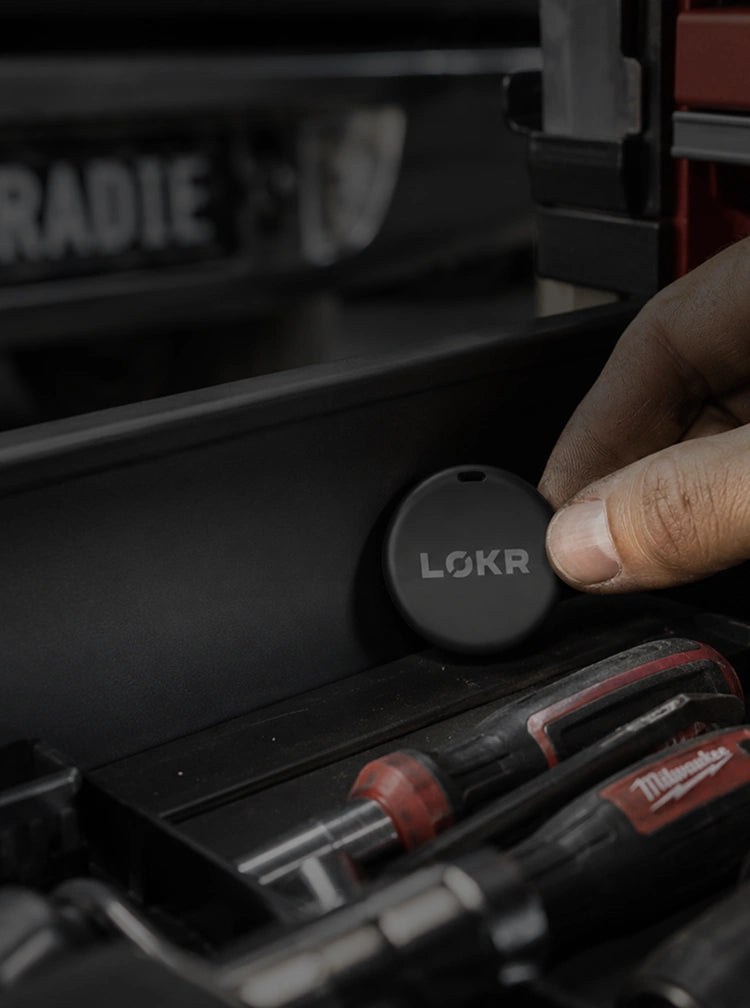 A close-up shot shows a hand placing a small, round, black tracking tag onto the inside wall of a dark-colored toolbox or drawer