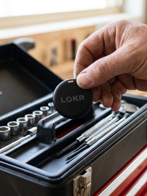A close-up view of a person's hand holding a LOKR tracking tag over an open toolbox filled with tools