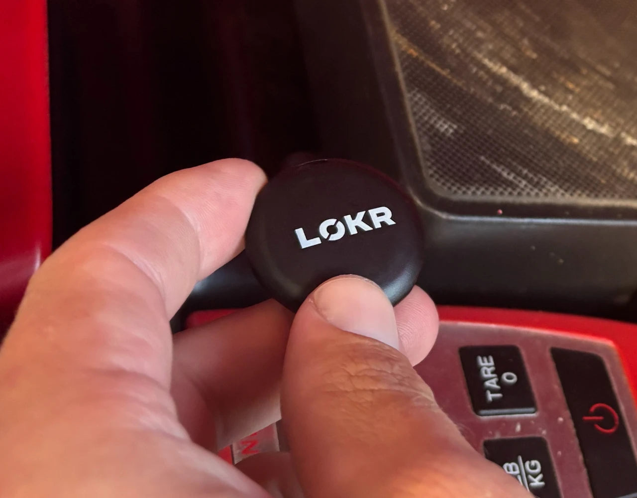 A close-up shot of a hand holding a LOKR tracking tag with white branding text, positioned above a digital scale